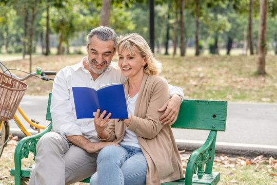Happy couple senior reading a book together while relaxing and sitting on the bench in the park