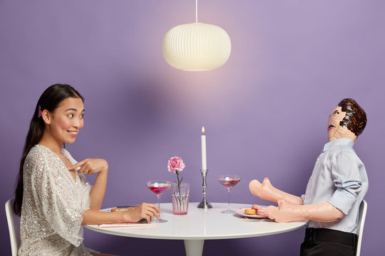 Pleased Asian woman sorts out relationships with layman doll, points at herself and asks why she should do first step, sits at festive round table opposite inflated unreal man. Relationship imitation