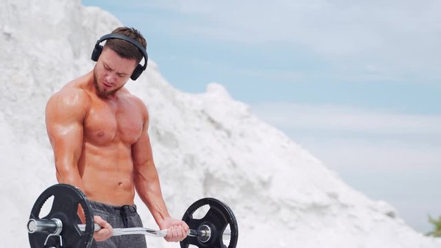 A man in headphones does barbell biceps exercises. Workout in nature.