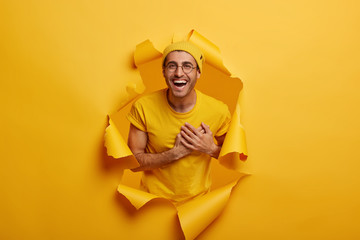 Satisfied man stands in paper hole, keeps hands near heart, expresses gratitude, being pleased by heartwarming compliment, stands in ripped paper hole of yellow background. Thankful cheerful guy