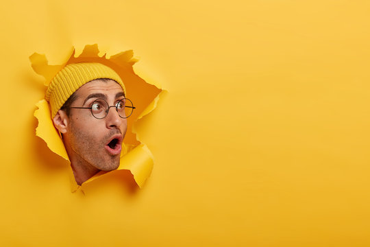 Stunned man looks with great surprisement or fear aside, opens mouth widely, wears hat and round glasses, looks through paper hole, isolated over yellow background. Shocking relevation concept