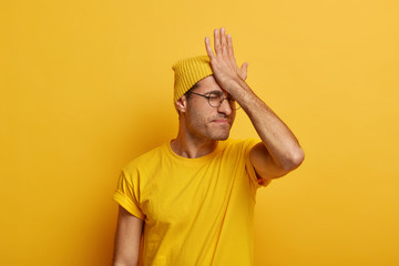 Serious man forgot about important meeting, punches palm on forehead, has regretting feelings, remembers about task, wears casual clothes, models against yellow background, looks bothered, worried