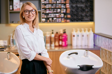 beautiful confident professional hairdresser in beauty saloon, caucasian woman with blond hair wearing white shirt stand looking at camera, isolated in beauty saloon