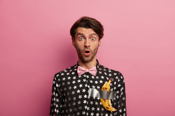 Conceptual photo of shocked man stares shocked at camera, wears pink bowtie, fashionable black shirt with printed stars and taped banana trend after visitng art gallery, impressed by artwork