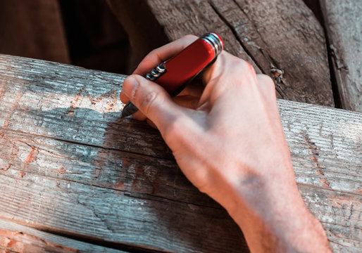 Hand holding a red multitool pocket knife carving a signature in a wooden beam. Act of vandalism