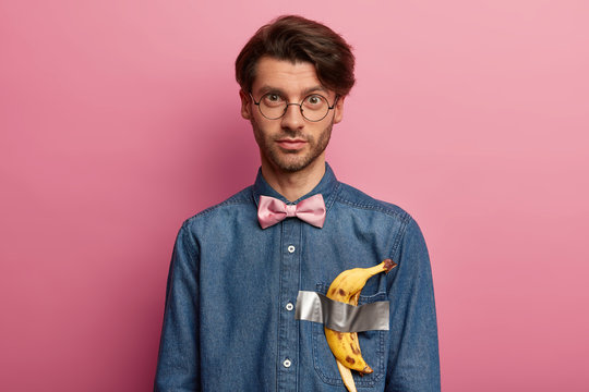 Imitating most popular and expensive piece of art. Handsome man wears clothes with banana duct tape, appreciates conceptual art, looks straightly at camera through glasses, stands over rosy wall