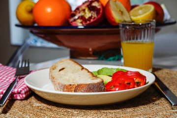 A piece of bread with avocado and peppers on a plate, with a ginger shot, and a fruit basket.