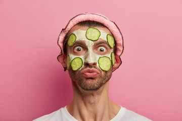 Face care concept. Shocked emotive man stares with bugged eyes at his reflection, applies white clay mask with slices of cucumber, wants to have natural beauty, proper approach to clean skin