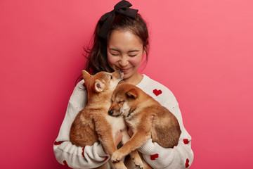 Sincere happy woman plays with two puppies, gets kiss from shiba inu dog, expresses love to...