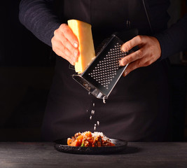 A man in an apron rubs cheese on a grater, prepares a dish. Cheese falls on food. Dark background.