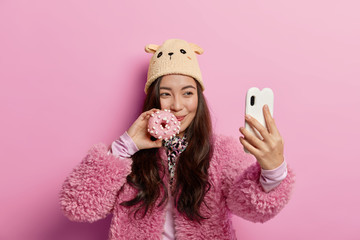 Pretty happy Korean girl poses with freshly baked doughnut, takes selfie portrait, unhealthy eating, shares photos in social networks, going to taste sweeties glazed donut, wears winter outerwear