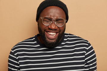 Headshot of dark skinned bearded man laughs out happily, squints from joy, shows white teeth, wears black hat and striped sweater, isolated over brown background, hears hilarious joke, has fun