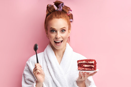 Young redhead woman wearing bathrobe, curlers after shower eating sweet tasty cake. In one hand spoon, in another red cake. Emotional girl posing with cake isolated over pink background