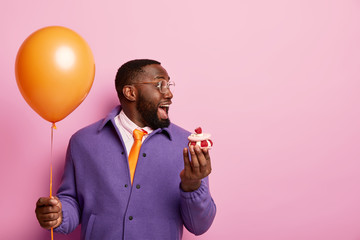 Cheerful shocked Afro American man stands with party balloon and sweet dessert, cannot believe eyes...