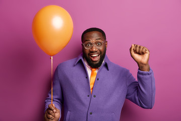 Horizontal shot of happy black man celebrates successful finishing of university or college, invites friends to share happiness, makes victory dance with clenched fist and inflated air balloon