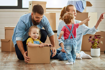 Caucasian family, man, woman and two girls play with moving box. Mother points to window of youngest daughter, father sat eldest daughter into box