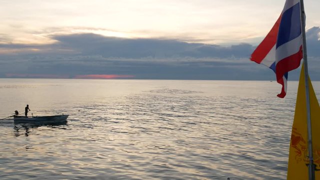 Boat floating near Thai flags in evening. Silhouette of anonymous person floating on boat on rippling sea near Thai and King of Thailand flags against cloudy sundown sky