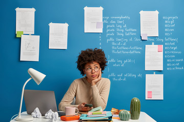 Shot of thoughtful female programmer holds mobile phone, thinks on publishing new post in social...