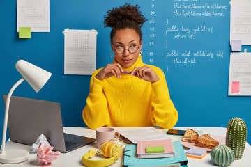 Contemplative dark skinned woman in yellow sweater holds pen, works as professional programmer, thinks about installing popular application from internet, has e learning, break for having snack