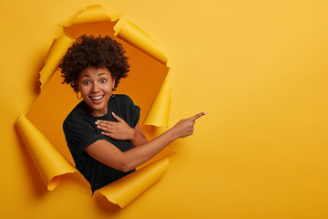 Happy curly haired African American woman laughs positively, points aside on copy space, wears...