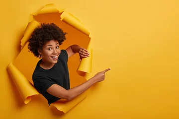 Poster Pleased dark skinned Afro American woman stands in ripped background, laughs happily, poses in paper hole, points on right side, isolated on yellow wall. Space for your advertising or promotion. © Wayhome Studio