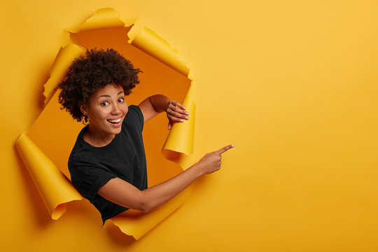 Pleased dark skinned Afro American woman stands in ripped background, laughs happily, poses in paper hole, points on right side, isolated on yellow wall. Space for your advertising or promotion.