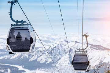 Cabin of a gondola cableway suspended on a rope where sits people with skis and snowboards high in the Altai mountains with snow and blue sky on winter sunset. Ski resorts and snowboarding.