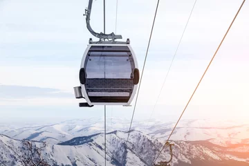 Garden poster Gondolas Cabin of a gondola cableway suspended on a rope where sits people with skis and snowboards high in the Altai mountains with snow and blue sky on winter sunset. Ski resorts and snowboarding.