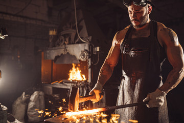 portrait of blacksmith in leather apron holding hot metal, wearing brown leather apron, space with furnace, fire - Powered by Adobe