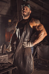 Portrait of serious confident strong young metalworker in black apron stand looking at camera, work...
