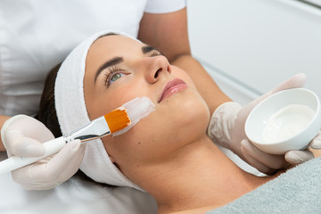Kosmetolog wykonuje zabieg pielęgnacyjny cery. Nakłada krem na twarz kobiecie w salonie...
