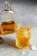 A glass of whiskey with ice and a half empty bottle on a gray concrete background. Selective focus.