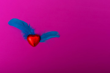 Red heart shape of chocolate arrange with blue and purple feathers on the pink background. The heart is flying and has wings. Copy space. Flat lay.