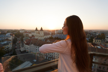 Fototapeta na wymiar Young beautiful woman admiring panoramic view of european street architecture high from the balcony. Girl looking at street panorama on sunset.