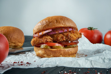 Tasty and juicy burger with pickled cucumbers, tomatoes, fried chicken and sweet onions on parchment. Fast food, street food.