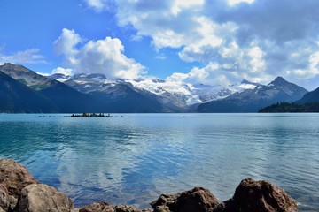 Beautiful blue Garibaldi Lake surrounded by high mountains with glaciers. Mysterious white and gray clouds, blue sky. An Island with stone pyramids. Rocky lake bank.