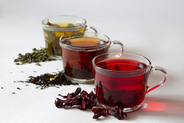Three glass cups with black, red and green tea with dry leaves on a white background. Red hot Hibiscus tea with dry flowers. Healthy lifestyle.  Green hot tea. Black hot tea. Hot drinks.