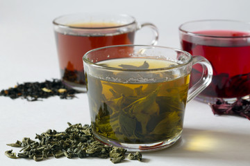 Green hot tea with dry leaves on a white table. Three glass cups with black, red and green tea on a white background. Red hot Hibiscus tea. Black hot tea. Healthy lifestyle. Hot drinks.