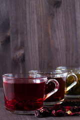 Red hot Hibiscus tea. Healthy lifestyle.Hot drinks.Three glass cups with black, red and green tea with dry leaves and flowers on a dark wooden background. Black hot tea. Green hot tea on wooden table.