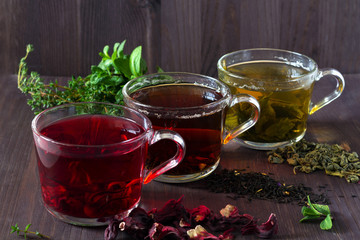 Healthy lifestyle. Three glass cups of black, red hibiscus and green herbal hot tea with dry leaves and flowers with fresh leaves of mint on a dark wooden background.Hot drinks.horizontal orientation.