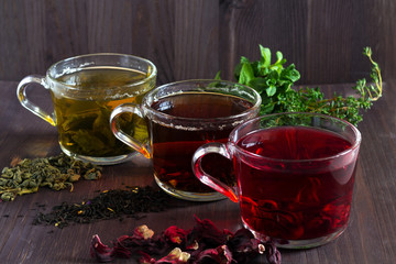 Three glass cups of black, red hibiscus and green herbal hot tea with dry leaves and flowers with fresh leaves of mint on a dark wooden background. Healthy lifestyle. Hot drinks. horizontal orientatio