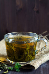 Glass cup of green herbal hot tea with dry leaves with fresh green leaves of mint with sackcloth on a dark wooden background. Hot drinks. Vertical orientation. copy space. Healthy lifestyle.