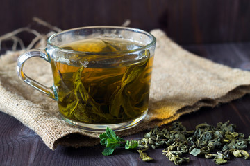 Glass cup of green herbal hot tea with dry leaves with fresh green leaves of mint with sackcloth on a dark wooden background. Hot drinks. Horizontal orientation. copy space. Healthy lifestyle.