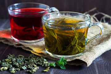 Hot drinks. Healthy lifestyle. Glass cup of green herbal hot tea and cup of red hibiscus tea with dry leaves with fresh green leaves of mint with sackcloth on a dark wooden background.