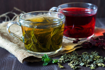 Healthy lifestyle. Glass cup of green herbal hot tea and cup of red hibiscus tea with dry leaves with fresh green leaves of mint with sackcloth on a dark wooden background. Hot drinks.