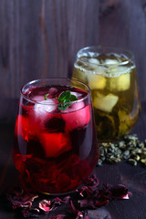 Healthy lifestyle. Glass of iced red hibiscus tea and dry flowers on a wooden table. Glass of iced green herbal tea with leaves of mint on a dark wooden background. Cold drinks. vertical orientation. 