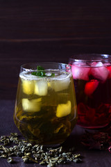 Glass of iced green herbal tea with leaves of mint on a dark wooden background. Healthy lifestyle. Glass of iced red hibiscus tea and dry flowers on a wooden table. Cold drinks. vertical orientation.