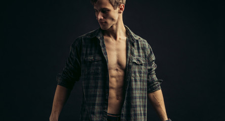 Portrait of a beautiful young athletic man in checkered shirt with opened naked torso in the studio, black background. Caucasian man looking side