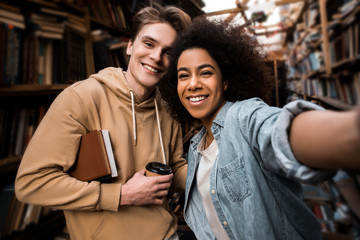 Selfie on phone, a multi-racial couple of students in the library are photographed in the library and rejoice in their achievements and productivity in learning. Education concept. Student lifestyle.