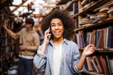 Happy student African-American woman talking phone, preparing for exams in university library, talkative and friendly concept, student lifestyle. Education concept